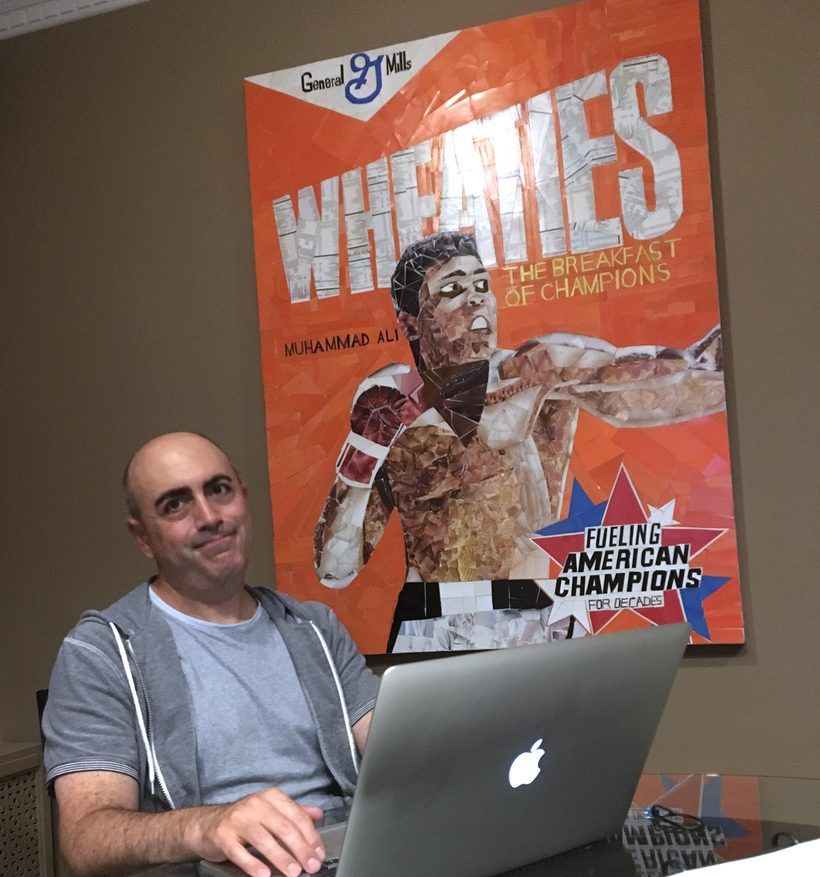 Greg working at a laptop with the finished Wheaties piece hanging behind him